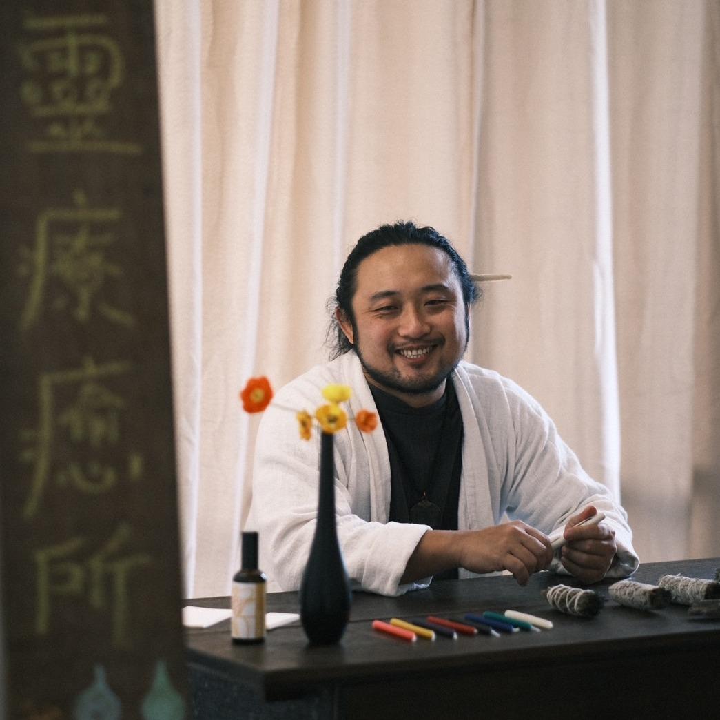 橘哥在問事現場的木招牌前微笑，桌上有乾燥花與蠟筆