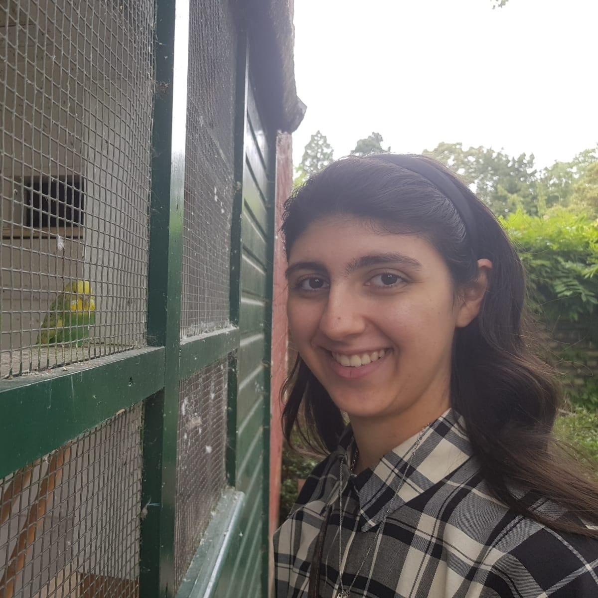 Picture shows Zahraa smiling. She is wearing a checkered black, grey and beige shirt. She is smiling next to a green budgie